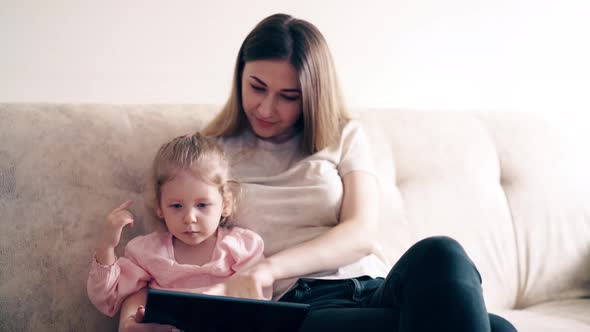 : Young Cute Mother and Little Girl Are Sitting on the Couch and Teaching Using a Tablet Computer alt