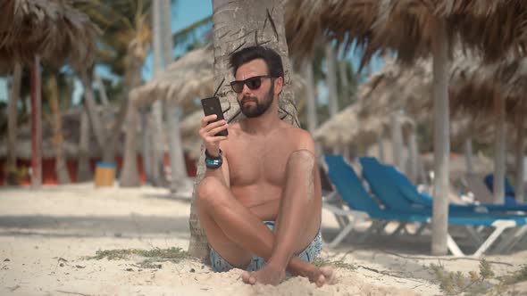 Man Sitting On Palm And Talking On Smartphone On Vacation Lounge Sea. Guy Relaxing On Tropical Beach alt
