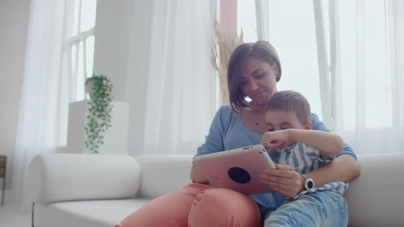 Mother And Son Using Tablet Looking At Screen And Talking On Sofa In Cozy House alt