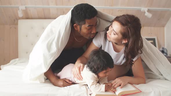 Happy Parents Reading a Book To Cheerful Child While Covering with a Blanket alt