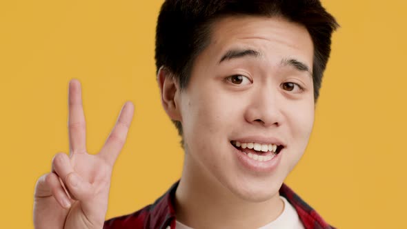 Cheerful Asian Guy Gesturing VSign Smiling Posing Over Yellow Background alt