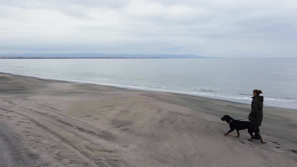 A Girl Walks with a Rottweiler Dog on a Leash Along the Beach in Cold Weather