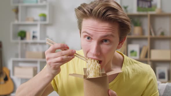 Young Man Eating Noodles From Box with Chopsticks at Home in the Living Room alt