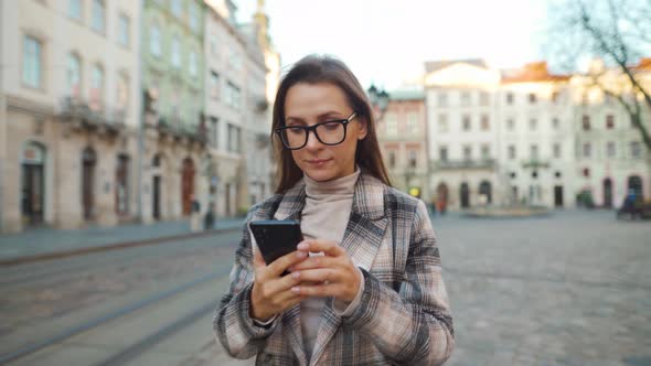 Woman Walking Down an Old Street, Using Smartphone and Taking a Photo alt