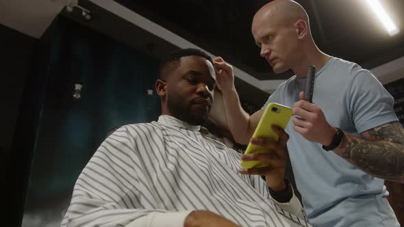 Bald Barber Combing the Hair of an African American Client Who Uses a Smartphone alt