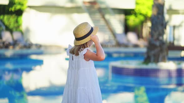 Cute Little Girl in a White Dress and Straw Hat By the Pool Near the Villa alt