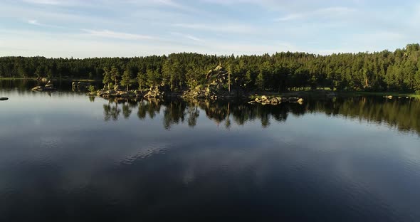 As the drone flies over a lake it reveals a man standing atop a rock cliff.  The water is smooth and alt