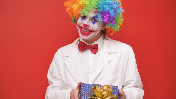 A Happy Clown Poses in Front of the Camera with a Gift Box on an Isolated Background