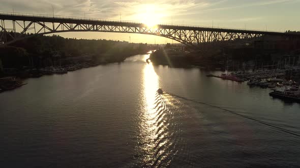 Aerial Following Boat Under City Bridge With Golden Sunset Flashing In Rafters