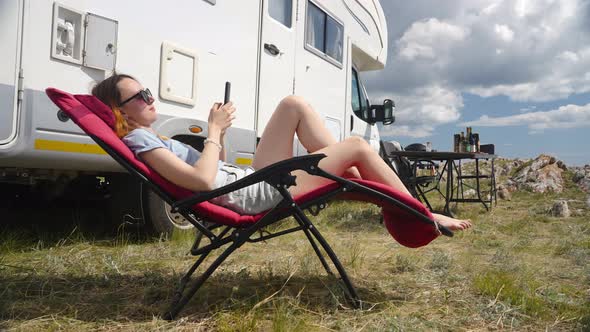 Woman Sitting On Folding Chair Using Sell Phone On Camp Site Next To Camper Rv alt