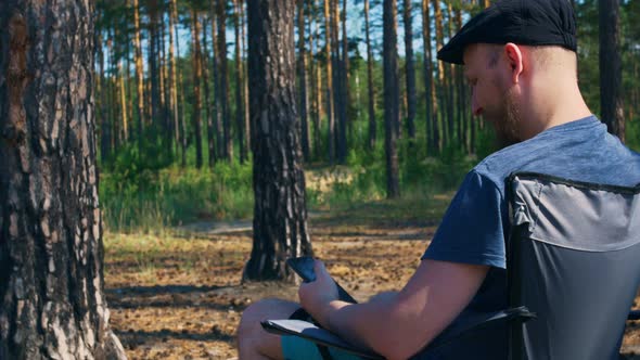 In a Clearing in the Forest a Young Man with a Wellgroomed Beard in a Summer Dress and a Cap Sits in alt