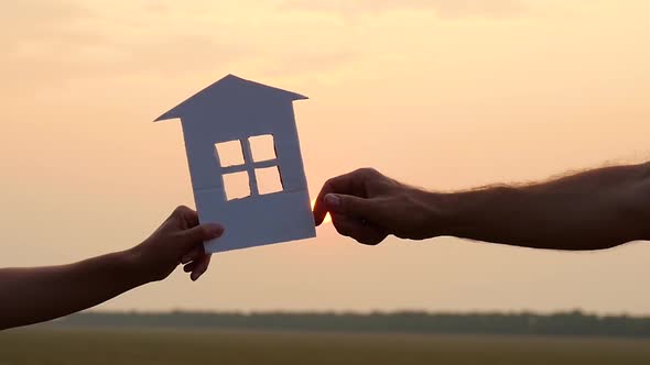A Man and a Woman Pass Each Other a Model of a Paper House on the Background of the Sunset. Concept alt