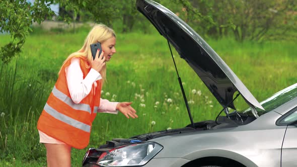 Young Attractive Blond Woman Wears Warning Vest and Phones with Smartphone in Front of Broken Car alt