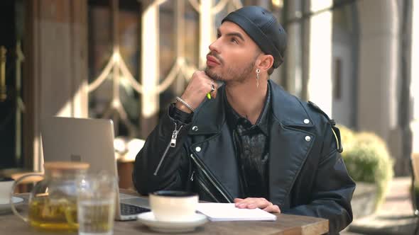 Thoughtful Handsome Gay Man Sitting in Sidewalk Cafe Writing Letter with Pen alt