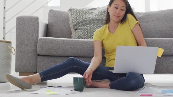 Focused asian woman sitting on floor, drinking coffee and working remotely from home with laptop alt