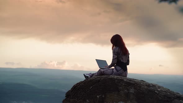 Girl Blogger Traveler is Typing Text on Laptop While Sitting on Top Mountain alt
