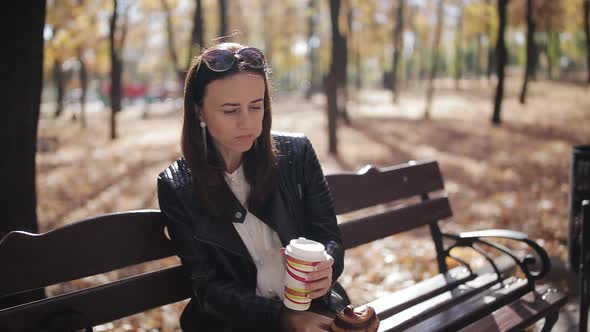 Business Girl on a Park Bench Eating a Croissant and Drinking Coffee From a Paper Cup alt