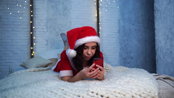 Young Woman in Santa Claus Costume Browses Smartphone Lying on Bed alt