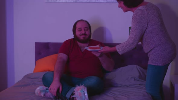 Young Fat Guy Watching Tv in Bed While Senior Mother Bringing Him Beer and Sandwich alt