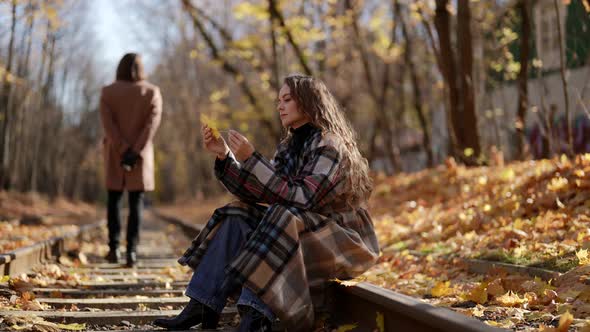 Woman is Sitting on Tram Rail in Autumn Day Playing with Leaf Silhouette of Man in Background alt