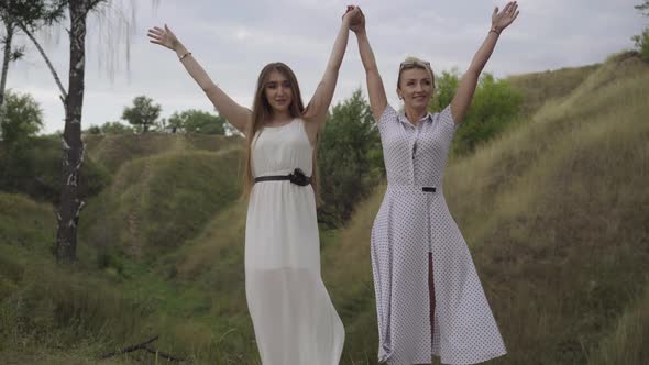 Wide Shot Portrait of Confident Gorgeous Caucasian Women in White Dresses Raising Hands on Overcast alt