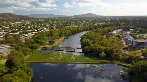 Drone flying above a calm reflective river with a dam wall/weir and a foot bridge, surrounded by hou alt
