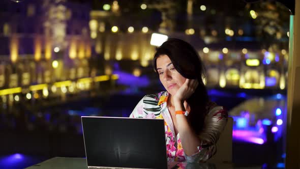 Beautiful Young Brunette Woman Uses Laptop, Sitting at Table on Open Terrace or Balcony, Against alt