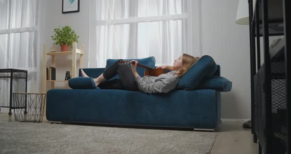 A Young Girl in Casual Clothes Lies Barefoot on a Sofa in a Modern Living Room alt