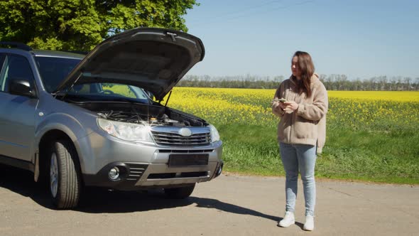 Woman Standing at Broken Car Searching for Repair Service on Smartphone alt