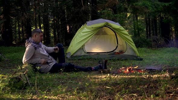 A Man Drinks Coffee Near a Tent in the Forest alt