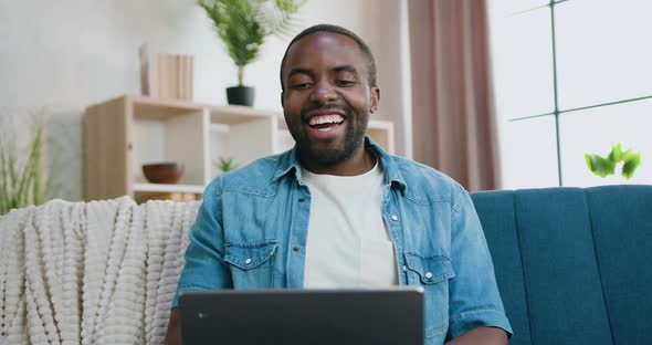 Bearded African Guy Sitting on the Soft Couch at Home During Pleasant Video Conversation on computer alt