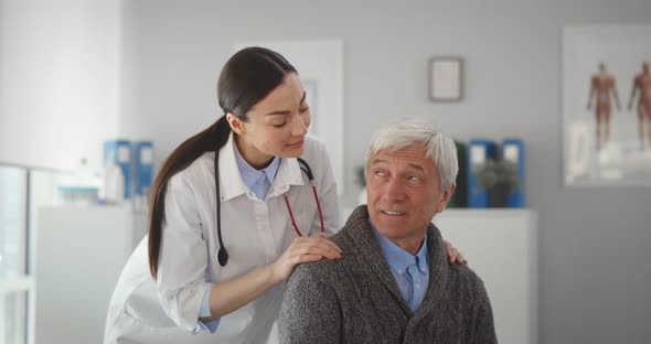 Female Doctor Hugging Smiling Senior Patient and Looking at Camera in Clinic Office alt