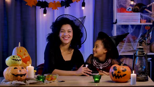 Mom and Daughter African American in Festive Costume and Witch Hat Joyfully Laugh and Have Fun alt
