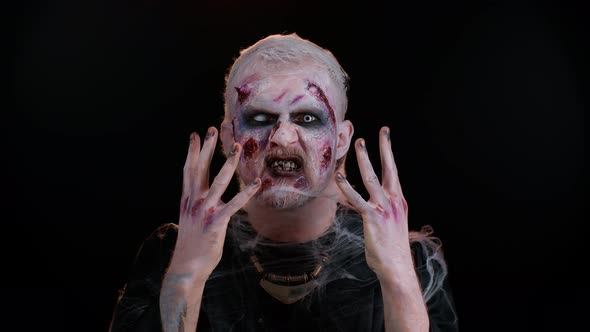 Scary Wounded Zombie Undead Guy Making Faces Looks at Camera Hiding Through Hands Smiles Terribly alt
