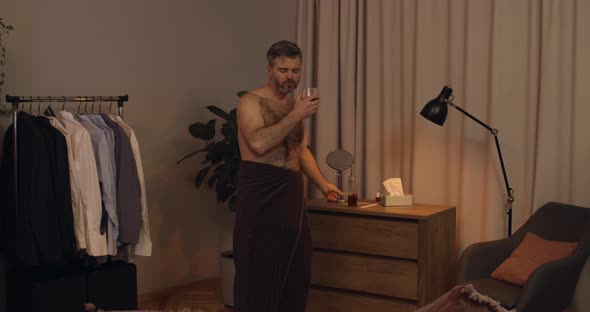 Handsome Bearded Guy in Bath Towel Pouring Elite Alcohol Into Glass and Drinking While Dancing alt