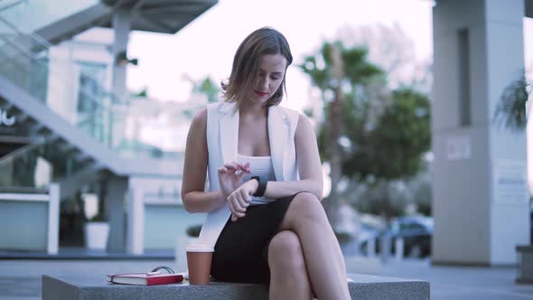 Young Stylish Woman Using Smartwatch While Sitting on Urban Bench