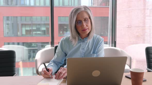 Senior Woman Works on Laptop and Making Notes in an Office alt
