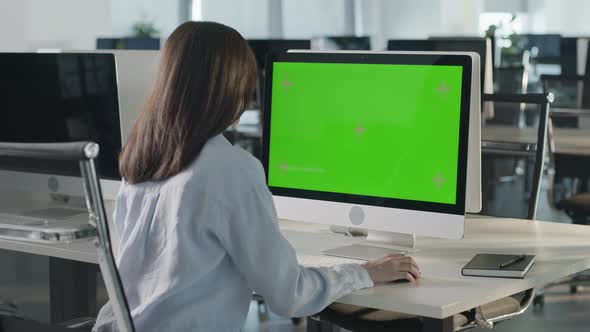 Asian Business Woman Typing on Desktop Computer with Green Mockup Screen While Sitting at His Desk alt