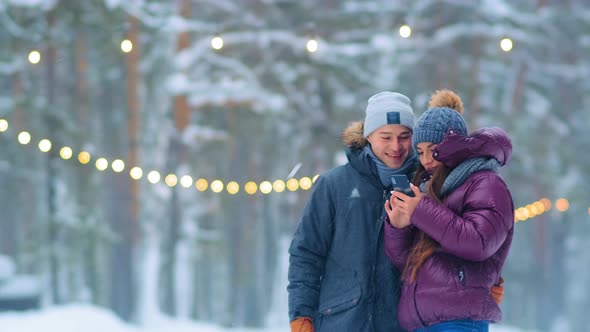 Young Couple in Warm Jackets Makes Selfie in Snowy City Park