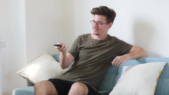 A Young European Man Is Sitting on the Couch Switching The Channel on the Remote Control That Does