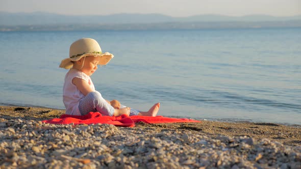 Lovely Baby Girl Sitting on Seashore. Little Tourist Sitting on Red Towel and Looking on Seascape