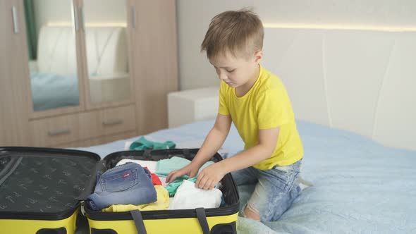 Cute Boy Sitting Near an Open Suitcase with Things for Vacation and Playing with a Selfie Stick for alt