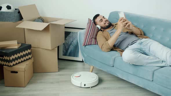 Carefree Arab Man Enjoying Smartphone and Music While Robotic Vacuum Cleaner Working in New alt