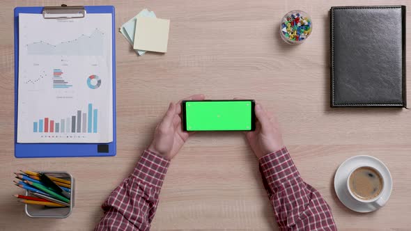 Man's Hands Over a Office Desk Holding Smart Phone with Green Screen alt