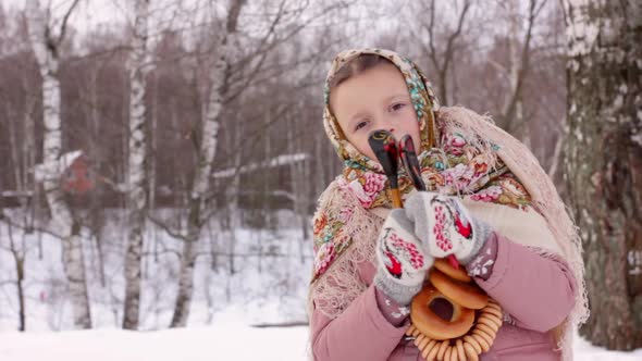 Cute girl in a traditional Russian headscarf and mittens playing on spoons on winter forest backgrou alt