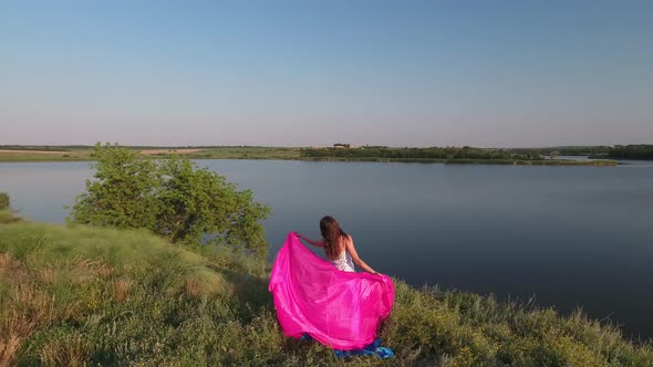 A Girl with Loose Hair in a Dress Stands on a Pink Cloth on the High Bank of the Lake