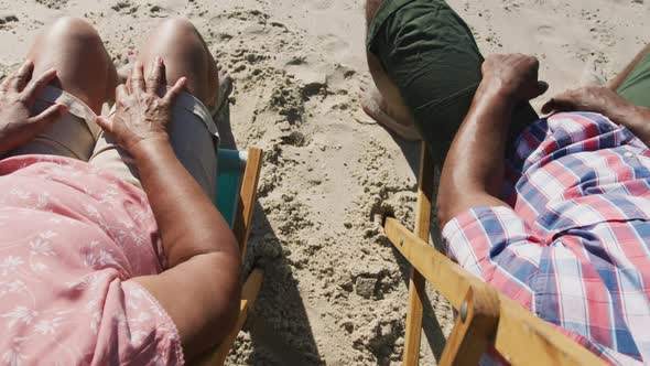 Senior african american couple sitting on sunbeds and holding hands at the beach alt