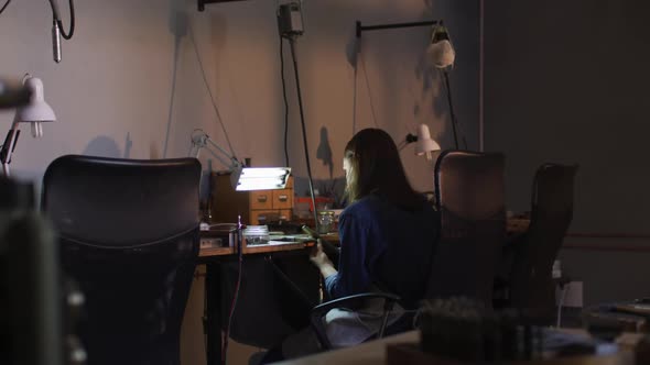 Back view of caucasian female jeweller sitting at desk, making jewelry in workshop alt
