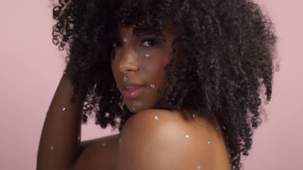Mixed Race Black Woman with Curly Hair Covered By Crystal Makeup on Pink Background in Studio alt