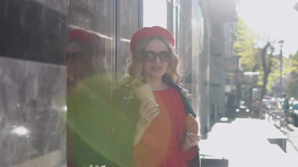Stylish Young Blonde in a Red Dress and Black Braid Stands on a Cozy Street in the Morning Sun alt